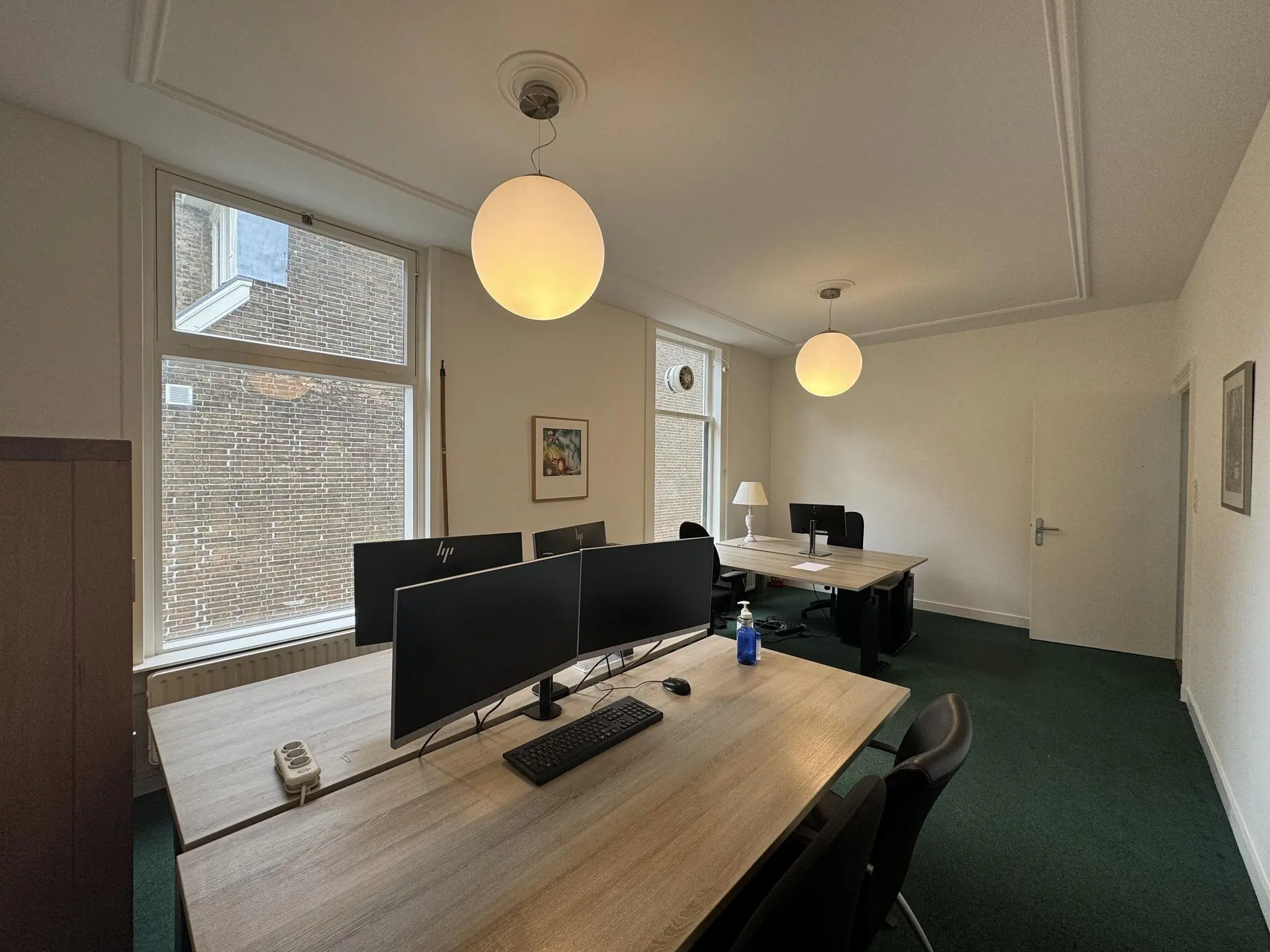 Kantoorruimte aan de Dorpsstraat met vier werkplekken, moderne bureaus en grote ramen met uitzicht op een bakstenen buitenmuur.